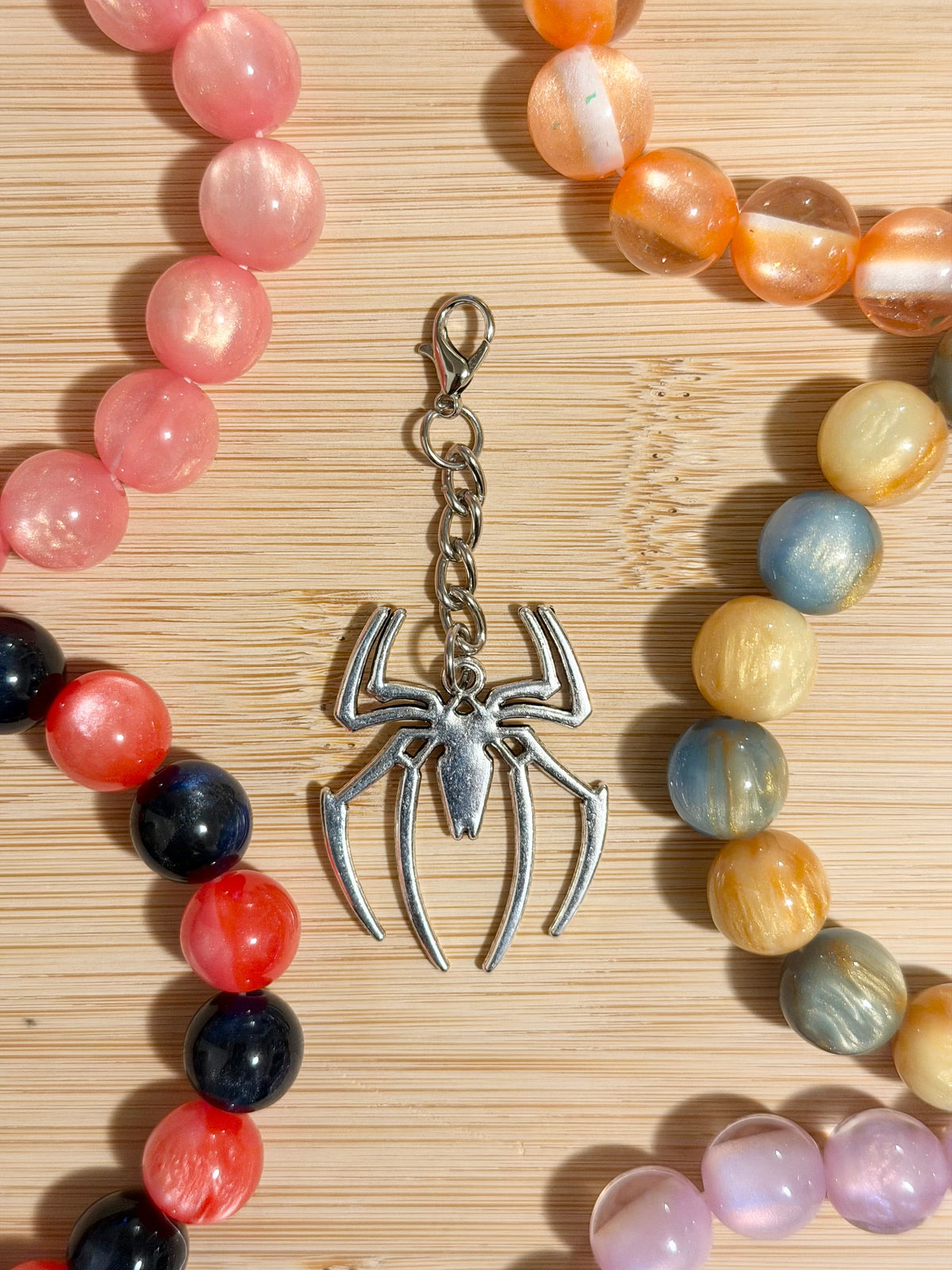 Large antique silver spider charm with long legs on a lobster clasp chain, surrounded by colorful beaded bracelets on a wooden surface