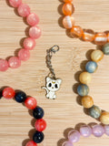White enamel chibi cat charm with gold crescent moon on forehead on a silver lobster clasp chain, surrounded by colorful beaded bracelets on a wooden surface