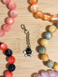 Black enamel skull and crossbones charm with red heart eyes on a silver lobster clasp chain, surrounded by colorful beaded bracelets on a wooden surface