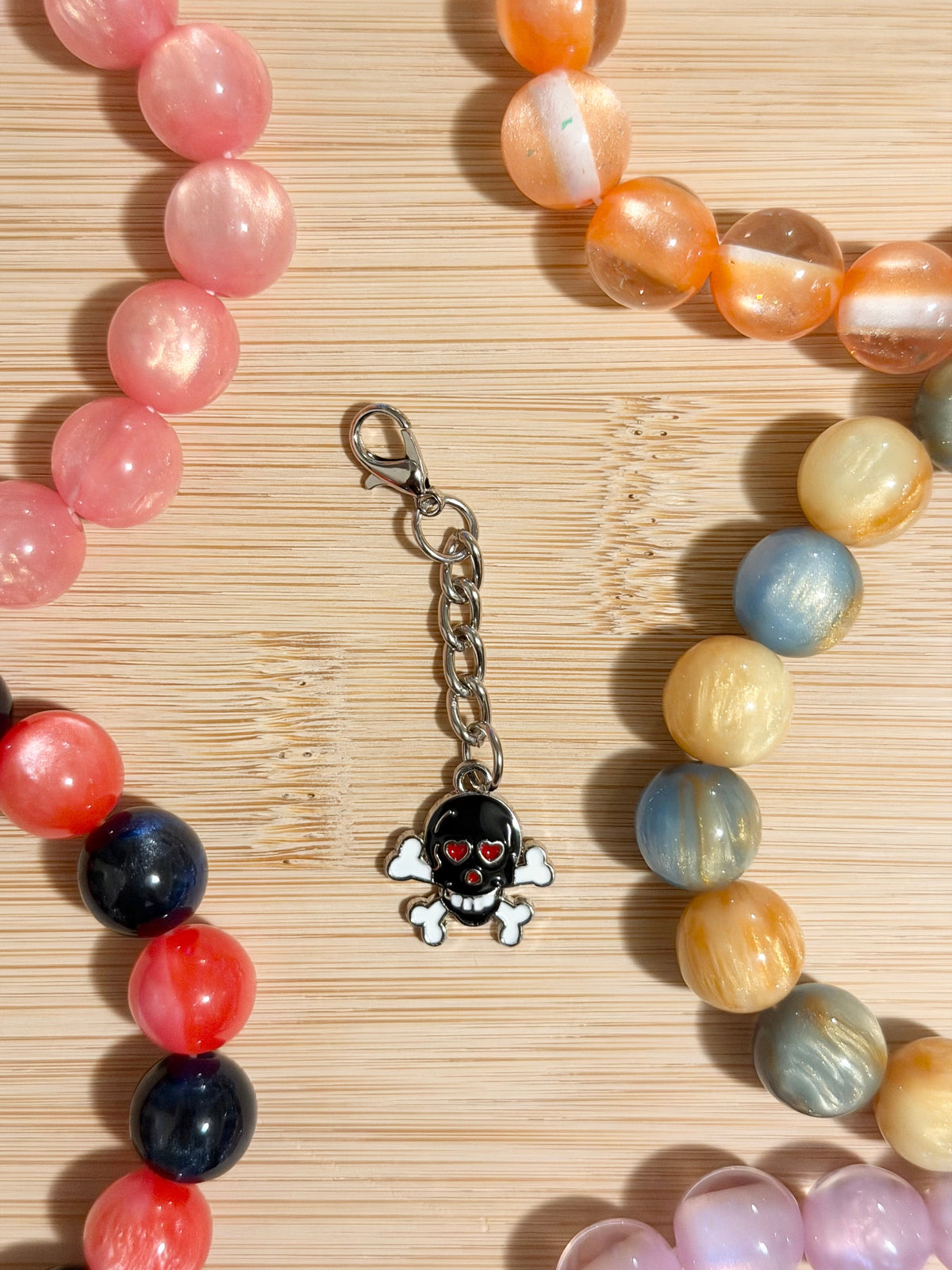 Black enamel skull and crossbones charm with red heart eyes on a silver lobster clasp chain, surrounded by colorful beaded bracelets on a wooden surface