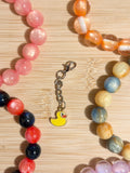 Yellow enamel rubber duck charm on a silver lobster clasp chain, surrounded by colorful beaded bracelets on a wooden surface