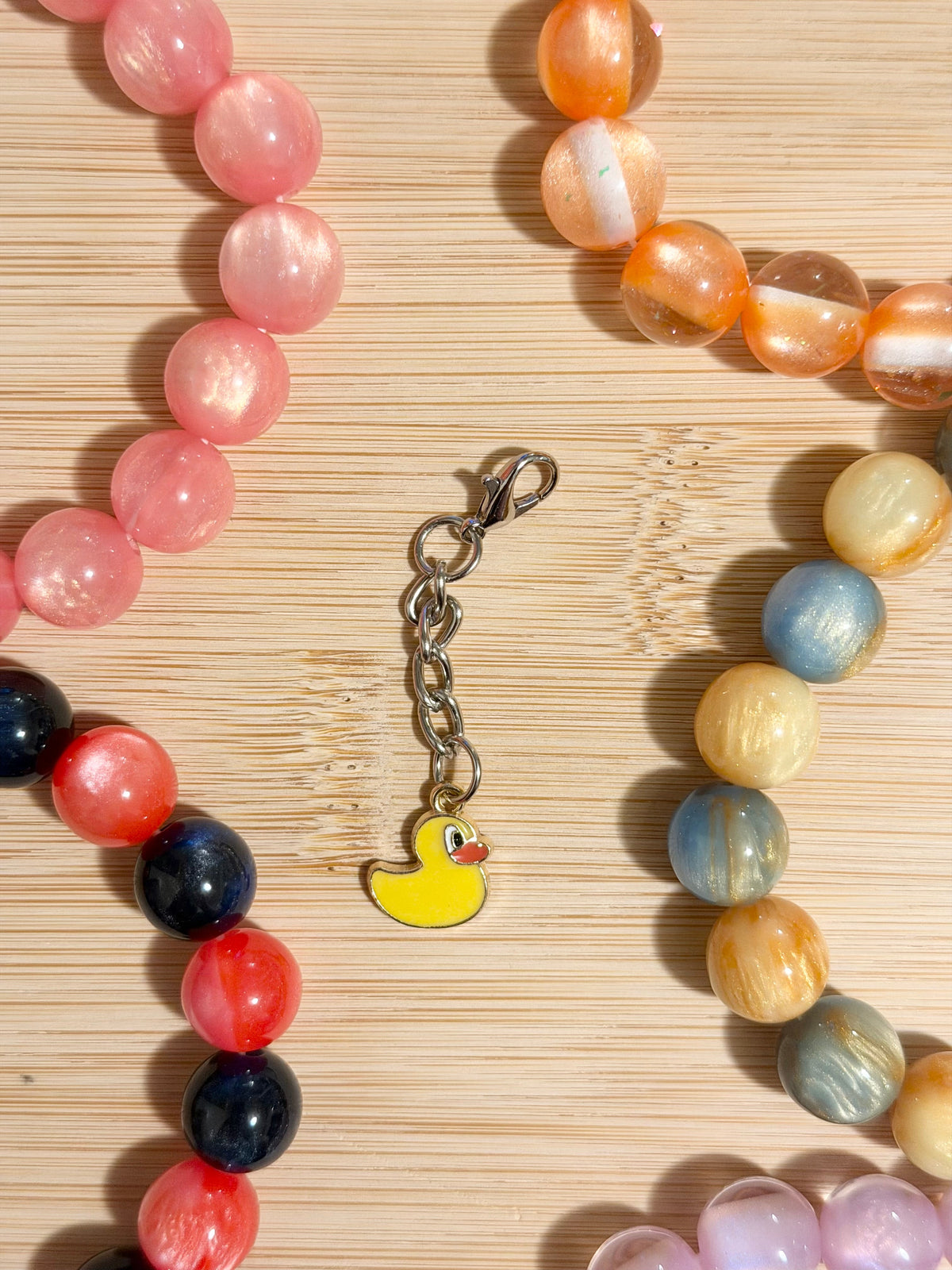 Yellow enamel rubber duck charm on a silver lobster clasp chain, surrounded by colorful beaded bracelets on a wooden surface