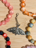 Large antique silver moth charm with detailed engraved wings on a lobster clasp chain, surrounded by colorful beaded bracelets on a wooden surface