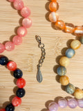 Delicate antique silver feather charm with engraved detailing on a lobster clasp chain, surrounded by colorful beaded bracelets on a wooden surface