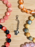 Antique silver sea turtle charm with detailed shell on a lobster clasp chain, surrounded by colorful beaded bracelets on a wooden surface