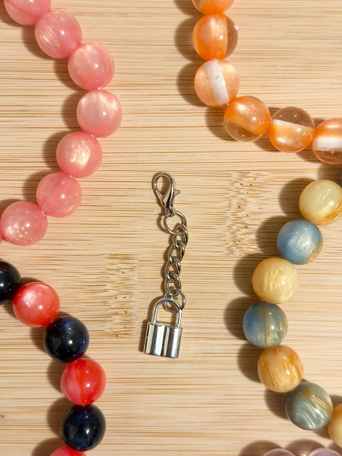 Shiny silver padlock charm on a lobster clasp chain, surrounded by colorful beaded bracelets on a wooden surface